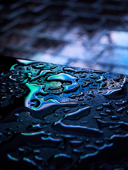 Blue-green neon lights are reflected in the rainwater pooled on the shiny surface of a black marble bench, while the out of focus paving stones in the background shimmer with pale blue light