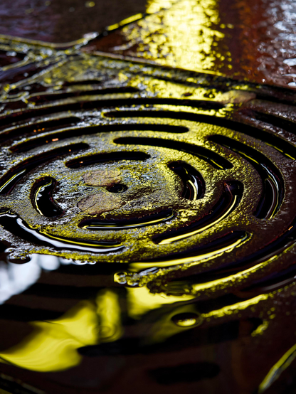 Golden light reflects on the rainwater around a circular iron street drain, making it look like it has been cast from precious metal