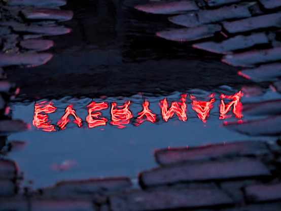 The word EVERYMAN, written in red neon capitals, reflects in a puddle among cobbled stones in an alleyway, the letters are reversed and shimmering because of a light breeze rippling the surface of the water