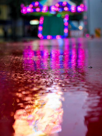 The metallic surface of a fairground ride reflects pink and blue neon lights in the background, along with the flame-like reflections from a neon advertising board in the foreground.