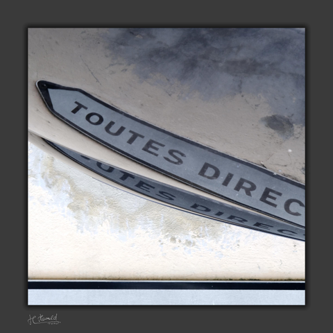 Square picture with dark grey frame. It shows the double reflection of a street sign reading ‘TOUTES DIREC’ in a bonnet in front of a light beige to grey wall. The sign is pointing to the top left. The image has been mirrored so that the text can be read correctly. There are a narrow black and a thick white horizontal lines at the bottom.
