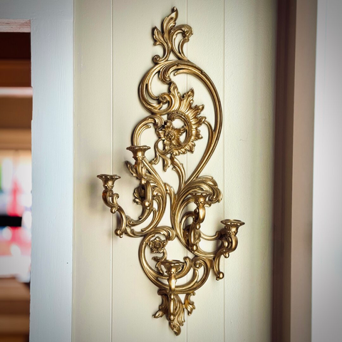 A thin, golden candelabrum hangs from a sand-colored wall next to a window through which morning light shines.