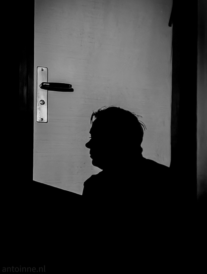 This is a high-contrast, black and white photograph that features a silhouette of a person in profile against a white door. The person's head and shoulders are visible, facing towards the left side of the frame. His hair appears somewhat disheveled. Behind and slightly to the left of the person's head is a metallic door handle, reflecting some light and standing out against the muted background. 