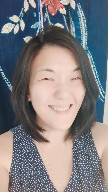 Photo of Joy Chew smiling and standing against an indigo blue background with flowers wearing a polka dot navy dress.