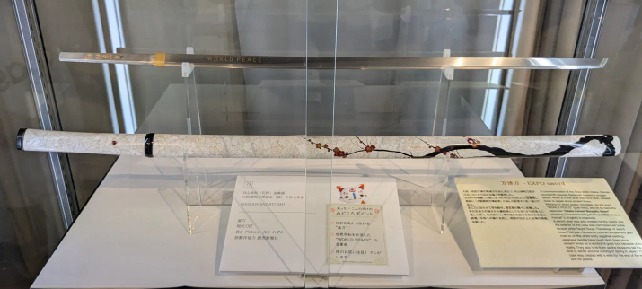 Dieses Schwert wurde von Gassan Sadatoshi gefertigt.
Oben hängt die Klinge unten der Halfter. Dieser ist überwiegend in weiss gehalten und mit einer Blumendeko bemalt. 
Unter dem Schwert liegen nähere Informationen in japanischer und englischer Sprache aus. 
Auf der Klinge steht in englisch "WORLD PEACE" geschrieben. 