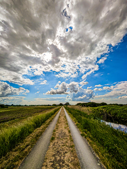 A road, flanked on both sides by fields and water, stretching into the distance. The sky dominates the upper portion of the photograph, filled with a mix of bright white, fluffy clouds and darker, more ominous gray clouds. The perspective of the road draws the viewer's eye towards the horizon, where a faint line of trees is visible. 