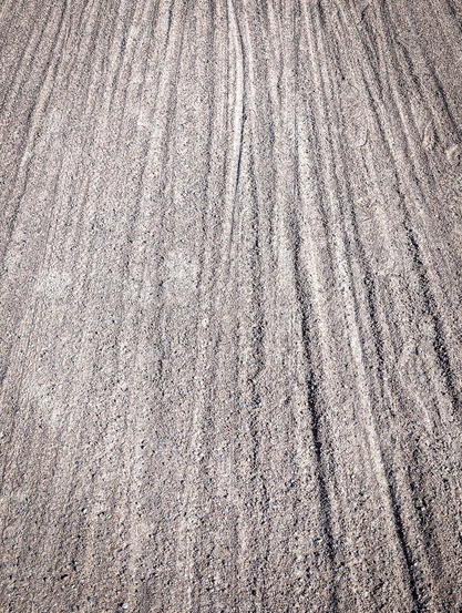 A gravelly surface with distinct linear patterns running vertically through it. The texture of the surface appears coarse, with individual grains visible. The lines, which appear to be indentations or ridges, are mostly parallel but show some slight variations in their straightness and depth. They seem to converge slightly towards the top of the frame, creating a sense of perspective and depth, as if looking down a path that recedes into the distance.