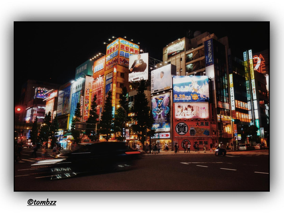 A nighttime urban street scene in Japan, bursting with vivid colors and dynamic energy.  
In the foreground, a blurred moving car adds a sense of motion and depth. Rising behind it are tall buildings covered in bright neon signs and advertisements, including colorful illustrations of anime characters and electronic store banners. Shades of blue, red, pink, and yellow dominate the scene, illuminating the night sky. At the base of the buildings, small silhouettes of people can be seen walking and crossing the street — bringing realism and emphasizing the lively nature of the location. Altogether, the image radiates the vibrant, neon-saturated buzz of a Japanese metropolis