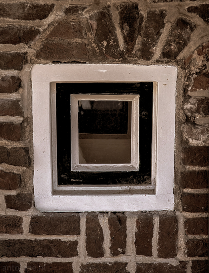 A square window set into a rustic brick wall. The window itself features a distinct black and white layered frame, creating an optical illusion of depth with a frame within a frame. The outermost frame is a thick, off-white or light beige, which stands out against the dark reddish-brown bricks. Inside this, there's a slightly smaller, dark frame. Within that, another, even smaller, off-white frame is visible, which then directly borders the actual glass of the window. 
