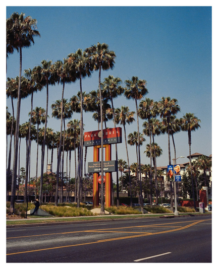 Rows of palm trees in descending heights. Underneath it is a sign saying “Park Brea”. On the left is one man in a wide-brimmed hat, leaning against a wall, and keeping himself cool under the palm’s shade. 