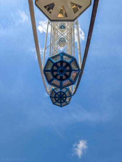 A frog's-eye view of a Ferris wheel against a bright blue sky with scattered white clouds. The perspective emphasizes the height and structure of the Ferris wheel, making it appear to extend upwards indefinitely. The Ferris wheel itself is predominantly light-colored, with a complex, geometric framework. Two blue, hexagonal cabins with multiple facets are visible, hanging below the main structure. 