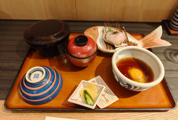 左上にご飯が入ったお櫃、右上に鯛の刺身、ほぼ中央の位置にお吸い物がある.
さらに、左下にご飯茶碗、右下に出汁と生卵の入った器があり、その中央下に漬物が入った小皿がある.