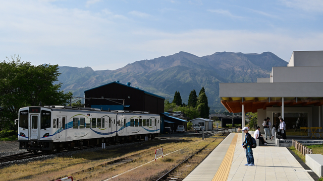 険しい山の麓に駅の車庫があり、列車が車庫側に留められている.
写真左側のクリーム色の列車には波のような模様が描かれている.
写真右側の駅ホームには、家族連れと少年が立っている.