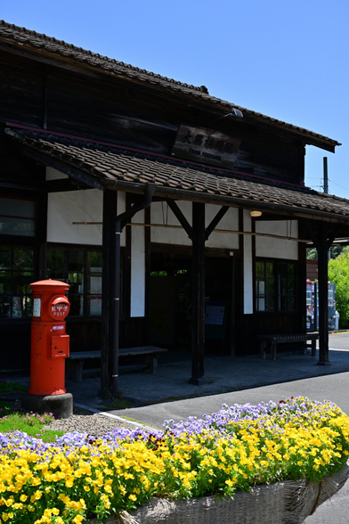 木造の駅舎が建っている.
駅舎入口左には丸くて赤いポスト、駅舎手前下には黄色い花の咲く花壇がある.