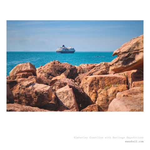 A little ship on the horizon, floating on stunning teal coloured waters with iron-oxide red rocks in the foreground.