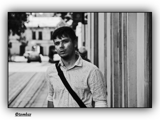 Black-and-white street photograph of a young man walking along an urban road. He’s wearing a long-sleeved striped shirt and has a shoulder strap from a bag crossing his torso. His hair is short, and his facial expression is calm and slightly contemplative. On the right side of the frame, there’s a smooth building wall. The background is softly blurred, revealing faint outlines of cars and city architecture.