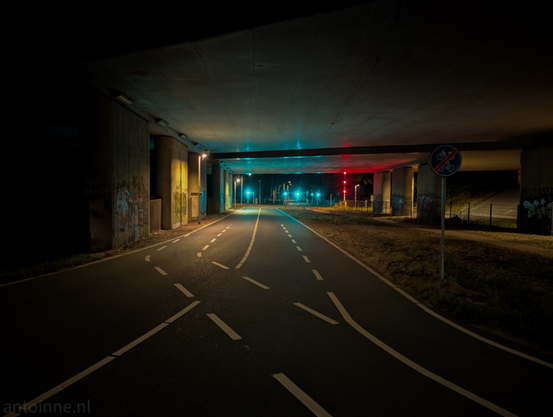 A night photograph looking down a paved road, as it passes underneath a concrete overpass. The scene is illuminated by artificial lights, creating a play of colors and shadows.

The pathway itself has white dashed lines indicating separate lanes, and it curves slightly to the left further down. On the left side of the pathway, there are vertical concrete supports or pillars of the overpass, some of which have graffiti. 

Streetlights with warm, yellow glows are visible along the left side, casting light onto the ground and the underside of the bridge. Further down the pathway, vibrant green and red lights are prominent. 