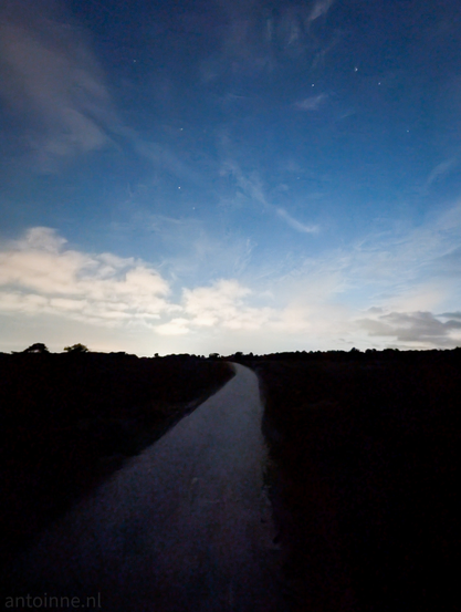 A low-light photograph of a path winding through a dark heathland landscape under a twilight sky. The prominent feature is a light-colored unpaved path that begins in the foreground and curves gently to the left as it recedes into the distance.

The path is illuminated by the ambient light of the sky. On either side of the path, the landscape is dark and silhouetted, Above, the sky transitions from a lighter, almost glowing horizon, to a deeper blue overhead. There are scattered clouds, particularly a band of brighter clouds near the horizon on the left, and wispy clouds higher up. Several stars are visible in the darker parts of the sky.