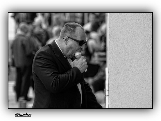 A man in a suit and dark sunglasses stands by a wall, partially tucked into the shadow. He’s focused on eating an ice cream, the gesture visibly expressive. The background features blurred figures, adding a sense of movement to the scene. The frame is distinctly split — a solid wall occupies the right side, contrasting with the open, lively space on the left.