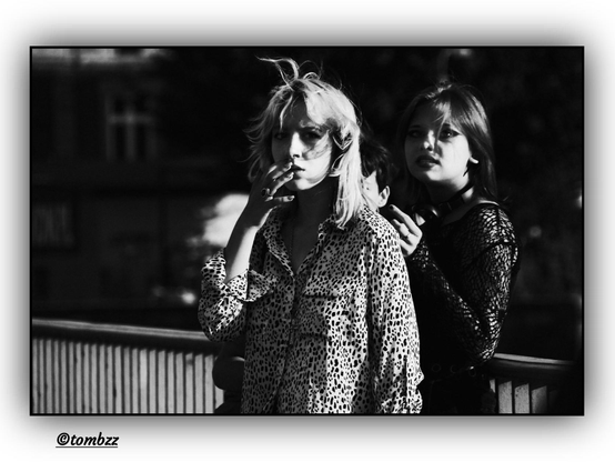 A black-and-white street photograph showing two young women standing side by side. The girl on the left has light, shoulder-length wavy hair, slightly tousled. Her face appears calm, almost contemplative. She’s wearing a light shirt with dark, irregular spots, resembling an animal-like pattern. Her left hand is raised gently to her lips — fingers loosely spread in a subtle, suspended gesture, adding emotional tension and intimacy to the shot, as if she’s lost in thought or mid-reflection. The girl on the right has long, straight dark hair, falling freely around her face. Her expression is more assertive, with a slight half-smile. She’s dressed in a dark, long-sleeved lace top. Behind them is a metal railing, and in the background stands a blurred building with faint window outlines, giving the photograph depth and an urban atmosphere.