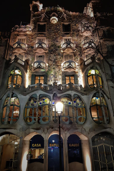 A striking, low-angle night shot of Casa Batlló in Barcelona, illuminated by warm, artificial light. The building's unique, organic facade is visible, with its skull-like balconies and colourful, mosaic-like detailing. The large, eye-shaped windows on the lower floors glow with interior light, revealing glimpses of chandeliers. In front of the building, a street lamp stands tall, casting a warm glow. The words "CASA BATLLÓ" and "GAUDI" are visible on the building's ground floor.