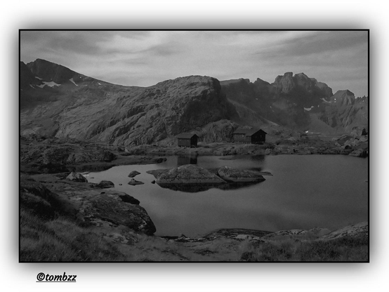 A monochrome mountain landscape featuring a tranquil lake in the foreground, surrounded by massive, dark boulders. Two wooden cabins with simple structures stand on the lake’s edge, adding a touch of rustic charm to the scene. In the background, majestic, rocky mountains rise, their peaks dusted with light snow, creating a striking contrast with the dark stone. Above the horizon, a partly clouded sky casts a melancholic and mysterious atmosphere. The image is rendered in shades of black, white, and gray, emphasizing the landscape’s drama and texture.
