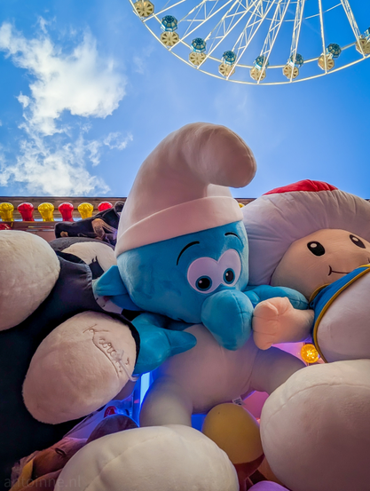 A Smurf plush toy surrounded by other stuffed animals, at a carnival or fair, with a Ferris wheel and a bright blue sky with clouds in the background.

The central focus is a large, light blue Smurf plush, easily identifiable by its white conical hat and friendly expression with wide black eyes. Above the plush toys, a significant portion of a Ferris wheel is visible against a vibrant blue sky with scattered white clouds, reinforcing the carnival setting. 