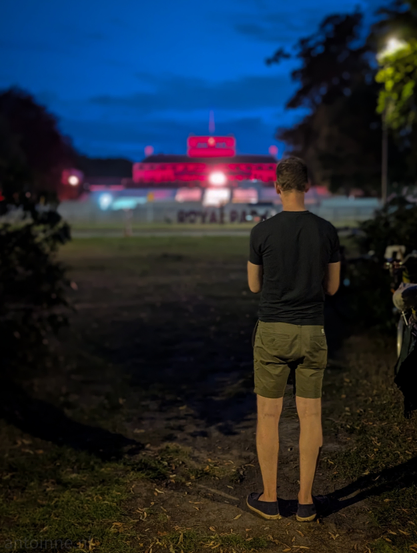 A man standing on a dirt path on the edge of an open grassy area, looking towards a building lit up in red in the distance. He is wearing a dark t-shirt, light-colored shorthand dark shoes. His back is to the camera, and his gaze is directed towards the brightly illuminated building. The palace (Soestdijk) in the background is prominently lit with a vibrant red light, standing out against the dark blue twilight sky. There are some white structures in front of the building, marked with "ROYAL PARK".