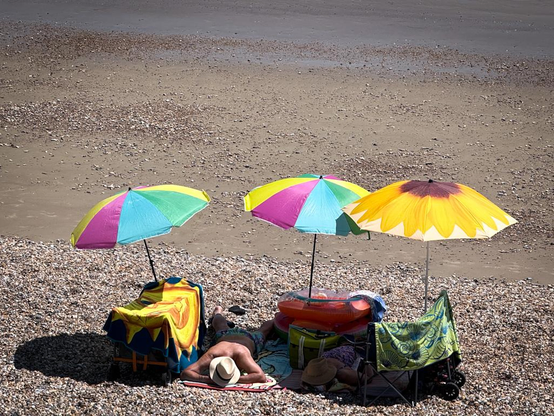 A man dressed only in swimming trunks and a white straw fedora hat is lying on his stomach on a blanket on a pebble beach next to a woman in a purple floral dress and a similar hat, lying on her back with her head on a cushion. There is a beach trolley to the left of the man that is covered in a blue and yellow rug, and a pile made up of two small plastic inflatables and a large green tote bag between the couple. There is also what looks like a baby buggy to the right of the woman covered in a green towel with a number of blue shell motifs on it. Two sunshade umbrellas with rainbow coloured segments are attached to the top of the trolley and the inflatables, and a third umbrella with a sunflower motif is on the top of the baby buggy.
