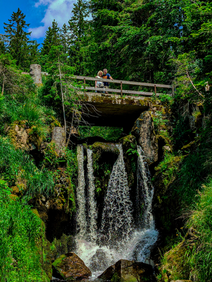 A picturesque natural scene with a couple enjoying the view from a bridge over a cascading waterfall. In the foreground, a multi-tiered waterfall dominates the lower half of the image, with white, frothy water dramatically splashing over dark rocks into a pool below. The rocks around the waterfall are covered in lush green moss and foliage, indicating a damp and fertile environment. The background is filled with dense, tall evergreen trees with a dark green coniferous forest. The sky is visible in the upper left corner, showing patches of blue with some soft white clouds. 
