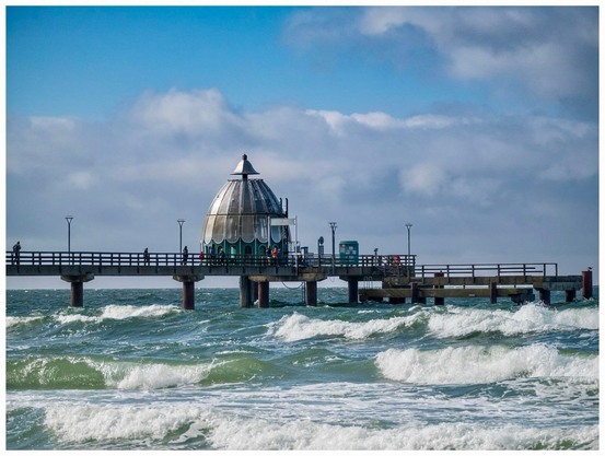 The seaside pier in Zingst (Baltic Sea) extends into the ocean, featuring a distinctive dome-shaped structure at the end. Waves crash against the pier, and the sky is partially cloudyy 