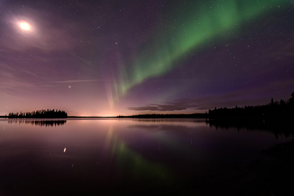 A long green ribbon of northern lights extends across a purplish, starry night sky from centre near the horizon up to the right corner. The moon is full and casting a flare in the top left corner; the lake below reflects back the lights and forest. A small forested island is visible on the left, and distant forest slopes up in silhouette on the right.