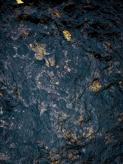 An abstract shot of water with visible elements beneath the surface. The water itself appears dark, almost black, but its surface is rippled and textured, creating patterns of light and shadow. There are numerous small, irregular ripples and waves across the entire frame. Beneath the dark water, there are lighter, irregular patches that appear to be rocks, pebbles and fallen leaves on the bed of the water body. These patches have a golden-brown or yellowish hue and are scattered throughout the darker water. Some of these lighter areas seem to reflect light, creating a shimmering effect.