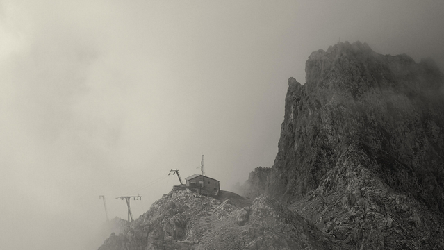 Das Foto zeigt eine dramatische Berglandschaft, die in Schwarz-Weiß gehalten ist. Im Vordergrund befindet sich ein steiler, felsiger Berghang, auf dem ein kleines Gebäude steht. Dieses Gebäude scheint eine Art Schutzhütte oder eine Station zu sein, möglicherweise für Wanderer oder Bergsteiger. Es ist über eine steile Treppe erreichbar, die sich den Hang hinaufschlängelt.

In der Nähe des Gebäudes sind einige mechanische Strukturen zu erkennen, die wie Teile einer Seilbahn oder eines Lifts aussehen. Diese Strukturen deuten darauf hin, dass das Gebäude möglicherweise eine Bergstation ist, die mit einer Seilbahn verbunden ist.

Im Hintergrund erhebt sich ein weiterer, noch höherer und steilerer Berg, der in dichten Nebel oder Wolken gehüllt ist. Die Wolken umhüllen die Spitze des Berges und verleihen der Szene eine mystische Atmosphäre. Die Sicht ist durch den Nebel eingeschränkt, was der Landschaft ein geheimnisvolles Aussehen verleiht.

Die gesamte Szene vermittelt ein Gefühl von Abgeschiedenheit und der rauen Schönheit der Natur in großer Höhe.