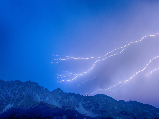 Es ist ein Gebirge im unteren Drittel des Bildes zu sehen, es handelt sich hierbei um die Nordkette. Über der Nordkette am nächtlichen Himmel sind (im Bild von rechts) 3 Blitze zu erkennen, die sich in viele kleinere Blitze verästeln. Der nächtliche Himmel erscheint durch die Blitze bläulich). 