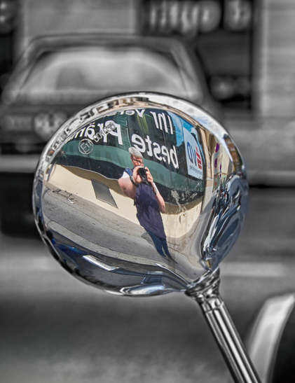 Ein Selfie auf der Rückseite eines Motorradspiegels. Ich stehe an einer Häuserecke, halte die Kamera halb vors Gesicht.  Die Glasfront der Häuserecke ist spiegelverkehrt beschriftet.
Der Hintergrund ist in schwarz-weiß gehalten. Man sieht den Teil eines Autos mit dem Heck zur Kameraansicht. Es ist eine weitere Häuserfront im Hintergrund zu sehen. Diese hat ein Fenster, dass spiegelverkehrt beschriftet ist.
In der Bildmitte sieht man eine Straße.