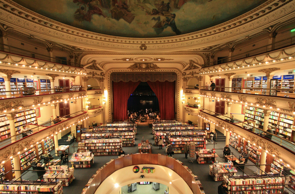 a beautiful and ornate theater has been converted into a bookstore, so instead of seating, there are many rows of books, though the red-curtained stage is still visible at the center of the photo