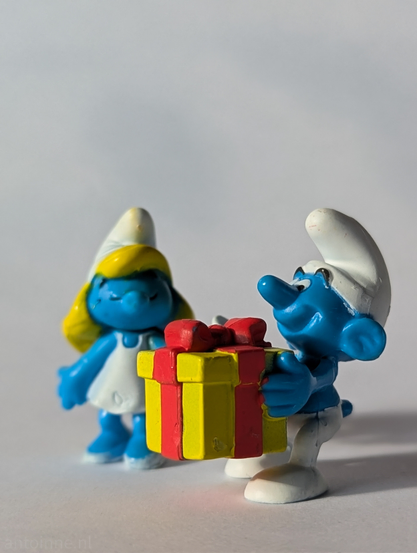 Two Smurf figurines on a plain, light-colored surface. In the foreground, slightly to the right, is a male Smurf holding a yellow gift box with a red ribbon and bow. He is facing slightly towards the left, looking at the gift. Behind him and to the left, slightly out of focus, is a female Smurf, wearing a white dress and with yellow hair. She is facing towards the male Smurf and the gift, and her eyes appear closed, suggesting a peaceful or happy expression. The overall impression is a joyful scene, depicting a  gift-giving occasion among the Smurfs. 
