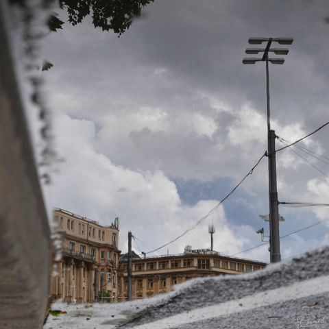 Square picture of a street scene reflected in a puddle. The image itself has been mirrored so that the buildings are not upside down. On the left, a ground stone is blurred in the picture and on the right is a tall lamp post with 6 lamps on top. There are many cables and wires hanging from it, as well as a surveillance camera. At the bottom right you can still see some pavement and above it (in the puddle) pale brown old multi-storey buildings on a street corner. There is an aerial mast on the house on the right.