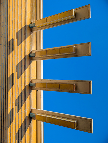Looking up at some of the lighting fixtures at the Hilversum Town Hall, designed by architect Willem Marinus Dudok. There are four horizontal elements. Each with a long, rectangular light fixture running along its underside.

They cast prominent, dark shadows onto the building's facade, creating diagonal patterns that contrast with the horizontal lines of the protrusions and the vertical lines of the building's texture.

The background is a vibrant, clear blue sky, free of clouds, which further emphasizes the architectural elements.