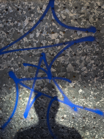 A dark, distinct shadow of a person's head and shoulders is cast upon the lower portion of a metallic surface, partially overlapping blue graffiti. The metallic, perforated surface, has a mottled, speckled appearance with varying shades of grey and silver. Small, dark, square indentations or protrusions are evenly spaced across the entire surface, creating a grid-like pattern.Strokes of vibrant blue graffiti, are sprayed haphazardly across the metal. These lines are thick and appear to form abstract, calligraphic shapes.
