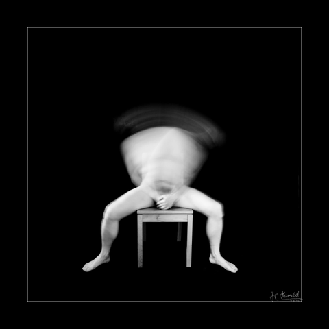 Square black and white picture with black frame of a naked male person sitting on a wooden chair. He moves his upper body to the right and left and a longer exposure blurs it. His hands cover his private parts.