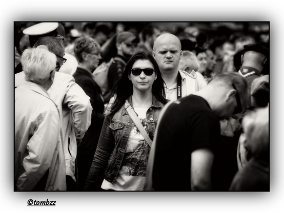 A black-and-white street photograph captures a crowd of people walking along a promenade. At the center of the frame stands a woman wearing large sunglasses, a denim jacket, and a printed T-shirt. She carries a light-colored bag over her shoulder, and her posture and expression convey confidence. Just behind her is a man in a light polo shirt, with a Canon camera hanging around his neck. His facial expression suggests curiosity or contemplation, as though he might be glancing at the woman walking ahead of him. The scene freezes a fleeting moment of everyday life, filled with subtle interactions and passing glances.