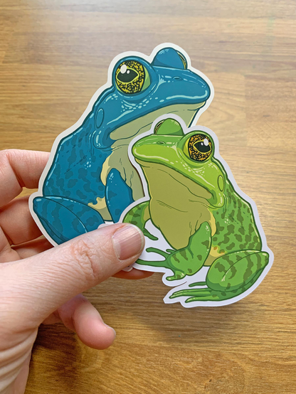 Photo of a hand holding a large sticker of a green bullfrog and an extra large sticker of a blue bullfrog