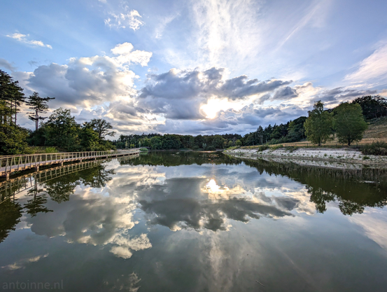 A wide-angle shot of a serene pond under a partly cloudy sky,  taken during the early evening. The sun is visible near the horizon, partially obscured by clouds, casting a bright reflection on the calm water. The clouds themselves are varied, with some fluffy white cumulus clouds and other more wispy cirrus clouds, creating a dynamic sky.

On the left side of the image, there's a wooden boardwalk or pier extending over the water. Lush green trees line the banks of the water body, with some taller, more pointed trees on the left and a more varied canopy on the right. The reflection of the trees and the sky, including the bright sun, is clearly visible in the still water, almost perfectly mirroring the scene above.