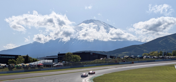 Zwei Rennwagen der SuperGT Serie durchfahren eine Linkskurve.
Im Hintergrund zeigt sich der Mount Fuji, leicht in ein Wolkenband gehüllt.