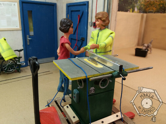The scene inside a 1:12 scale workshop. In the background are two blue doors, a Health and Safety at work act poster, a folded Brompton bicycle, a red dog lead hanging up, and an openroller shutter showing an enclosed yard outside the workshop.
There is also narrow gauge tram tracks set into the workshop floor.
The view outside is a work in progress.

Inside the workshop there are two Caucasian women doll figures. Harriet has dark hair, glasses, and is wearing a red tee shirt and blue jeans, Daisy has fair air and is wearing a grubby high viz coat. They are shaking hands.

In front of them is a large Wadkin AGS10 table saw painted green. It is on a wooden pallet and tied down with rope. The pallet is on an orange pallet truck having just been brought into the workshop through the roller shutter.

Outside in the yard is a grey and black needle felted mongrel dog called Monty Dog, and he is trying to pick up a black recycling collection box.

In the bottom right hand corner of the image is a watermark showing the Maker of Things logo consisting of an outline of a circular saw blade surrounded by a spanner, a hand saw, a ruler, a screwdriver, a cable and plug, and a pencil forming a hexagon. Inside the tools are the words Maker of Things.