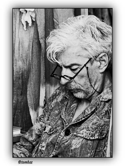 Black-and-white portrait of a man captured slightly from the side. He has gray, naturally styled hair and wears small, round glasses with thin frames. He’s dressed in a patterned shirt with a slightly sporty look. His head is tilted downward, and his gaze suggests he’s reading something. In the background, a curtain is clearly visible — textured and not uniform — adding a homely, intimate feel to the composition.