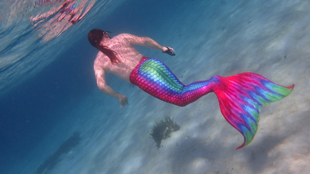 A merman with red hair swimming away. He has a red, blue, and green lower body and tail, and is swimming above a sandy sea floor.
