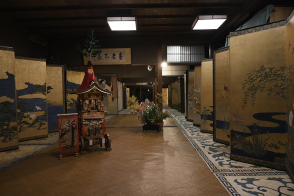 部屋の両脇に絨毯を敷いて、画面奥に向かって複数の便屏風が並べて飾られている。中央には北観音山のミニチュアと花も飾られている。/ 2025.07.22 新町通・六角町 藤井絞株式会社から